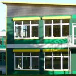 Eine Gesamtansicht von Außen zeigt das Schulgebäude in seiner Umgebung, in dunklem Grün gehüllt und mit gelben Akzenten versehen. Eine besondere Bauweise mit durchgeplanten Aufbau für den effektiven und schnellen Einsatz des Schulgebäudes.