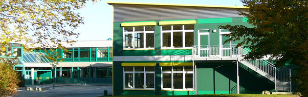 Eine Gesamtansicht von Außen zeigt das Schulgebäude in seiner Umgebung, in dunklem Grün gehüllt und mit gelben Akzenten versehen. Eine besondere Bauweise mit durchgeplanten Aufbau für den effektiven und schnellen Einsatz des Schulgebäudes.